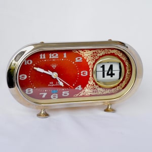 May include: Oval-shaped vintage clock with a red face and gold-colored trim. The clock displays the time and the date, with the number 14 visible. The clock has a gold-colored frame and small feet.
