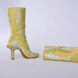 Può includere: Uno stivale a metà polpaccio con tacco alto e una pochette rettangolare abbinata. Entrambi gli articoli presentano un motivo ondulato e testurizzato nei toni del verde lime, giallo e argento, accentuato da piccoli cristalli riflettenti.