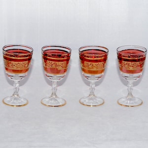 May include: Four vintage wine glasses filled with red liquid. The glasses have clear stems and bases, with gold rims and decorative gold bands around the bowls. They are arranged in a row on a white surface.