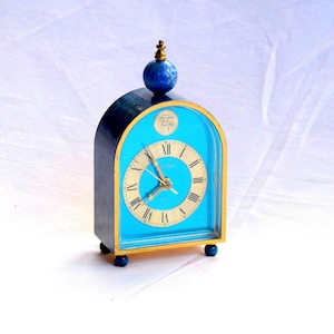 May include: A vintage blue and gold-toned clock with a round face and Roman numerals. The clock has a blue face and a gold-coloured frame. A blue sphere tops the clock. The clock is approximately 10 cm tall.