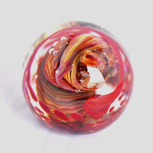 May include: A clear glass paperweight with a swirling design of red, orange, and brown colors. The paperweight has a round shape and small air bubbles are visible within the glass. The design resembles a fiery vortex.