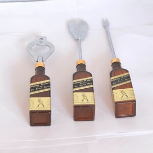 May include: Set of three bar tools with brown bottle-shaped handles featuring the "Johnnie Walker" label. The tools include a bottle opener, a spoon, and a fork, all with silver metal tops.