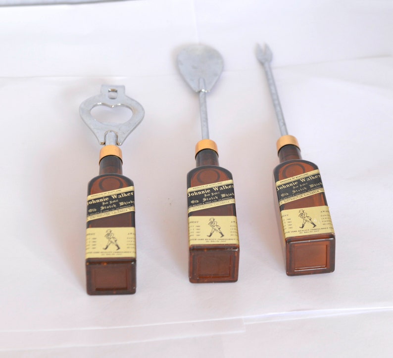 May include: Set of three bar tools with brown bottle-shaped handles featuring the "Johnnie Walker" label. The tools include a bottle opener, a spoon, and a fork, all with silver metal tops.