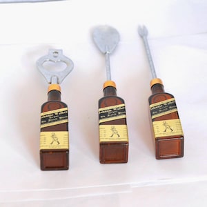May include: Three bar tools: a bottle opener, a spoon, and a fork. Each tool features a brown bottle base with a label that reads "Johnnie Walker". The metal tops are silver and the bottle tops are orange.