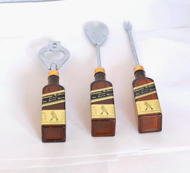 May include: Three bar tools: a bottle opener, a spoon, and a fork. Each tool features a brown bottle base with a label that reads "Johnnie Walker". The metal tops are silver and the bottle tops are orange.