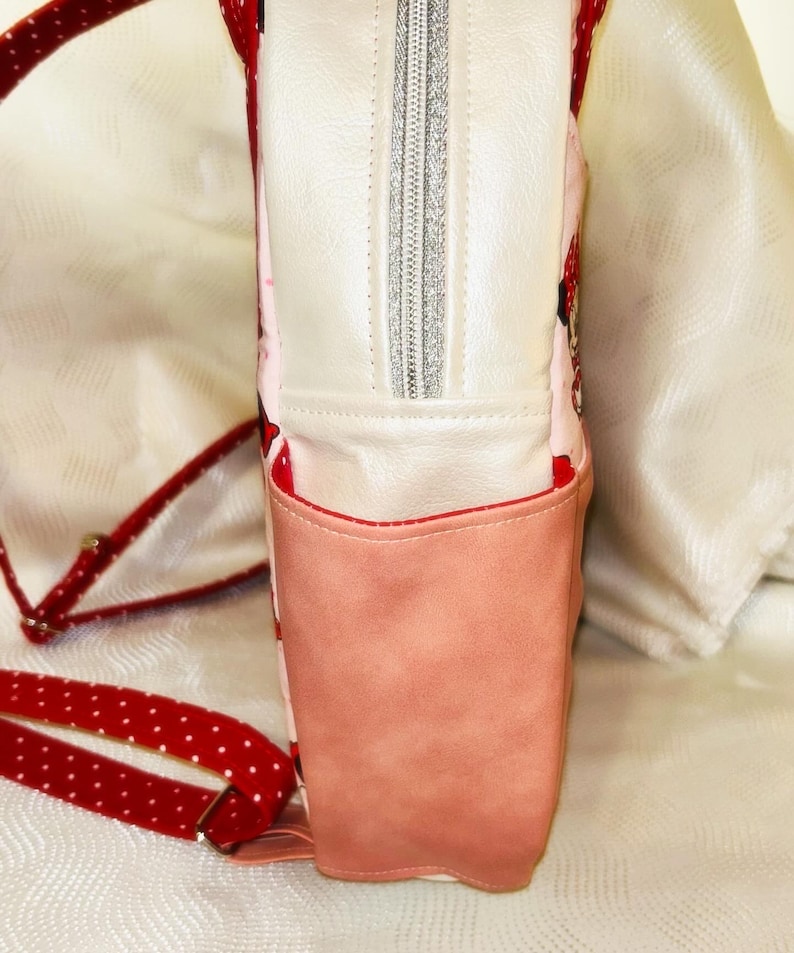 May include: A pink and white backpack with a red polka dot strap. The backpack has a front pocket and a silver zip. The interior has a pink pattern with red accents.