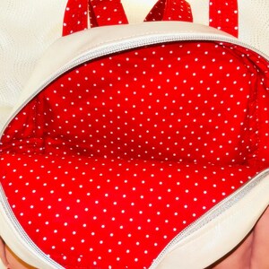 May include: Cream-coloured backpack with red polka dot lining and straps. The interior is a vibrant red with white polka dots. The zipper is silver. The straps are red with white polka dots.