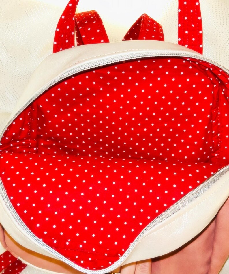 May include: Cream-coloured backpack with red polka dot lining and straps. The interior is a vibrant red with white polka dots. The zipper is silver. The straps are red with white polka dots.