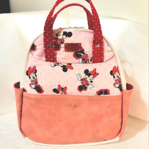 May include: A pink and white tote bag with a Minnie Mouse design. It has red polka dot handles and accents, and a peach-coloured front pocket. The bag is rounded and has a white base, suitable for carrying items.