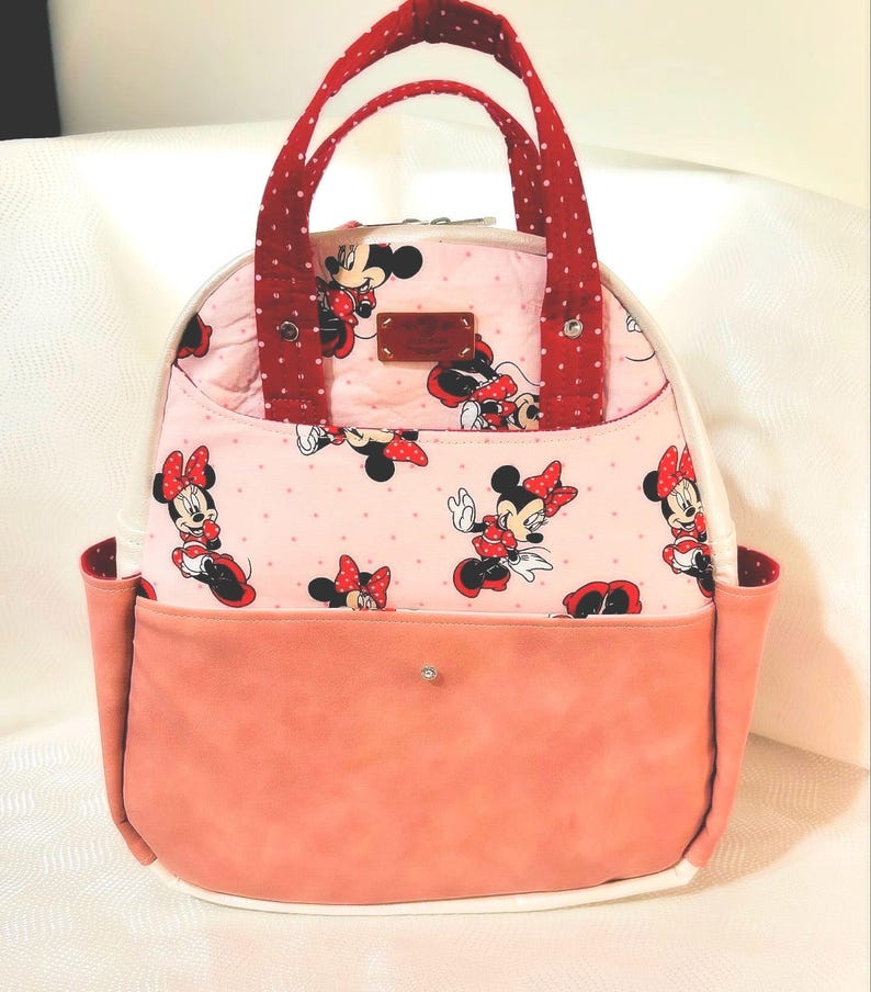 May include: A pink and white tote bag with a Minnie Mouse design. It has red polka dot handles and accents, and a peach-coloured front pocket. The bag is rounded and has a white base, suitable for carrying items.