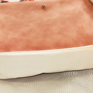 May include: A handbag with a light pink interior patterned with Minnie Mouse. The bag has a peach-coloured suede-like flap and a cream-coloured border. The background is a textured, silver-coloured surface.