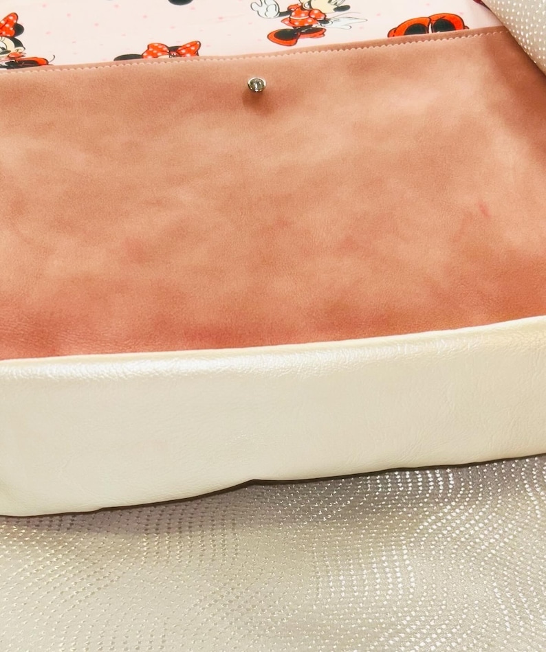 May include: A handbag with a light pink interior patterned with Minnie Mouse. The bag has a peach-coloured suede-like flap and a cream-coloured border. The background is a textured, silver-coloured surface.
