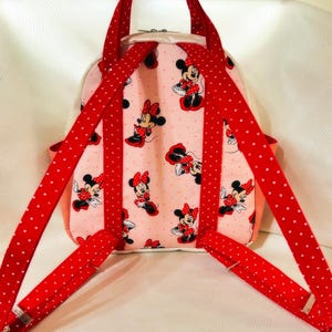 May include: Pink backpack with a Minnie Mouse pattern. It has red polka-dot straps and a handle. The bag features a zip closure and a light pink interior. The straps and handle are red with white polka dots.