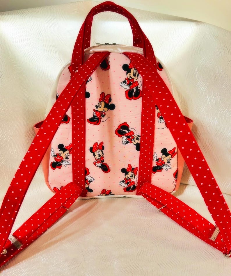 May include: Pink backpack with a Minnie Mouse pattern. It has red polka-dot straps and a handle. The bag features a zip closure and a light pink interior. The straps and handle are red with white polka dots.