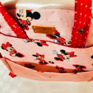 May include: A pink tote bag with a Minnie Mouse pattern. The bag has a light pink exterior, red polka dot straps, and a pink interior with a Minnie Mouse print. A small leather tag is visible.