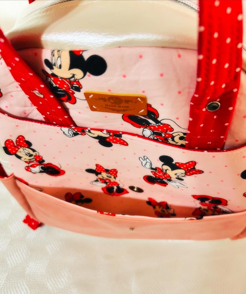 May include: A pink tote bag with a Minnie Mouse pattern. The bag has a light pink exterior, red polka dot straps, and a pink interior with a Minnie Mouse print. A small leather tag is visible.