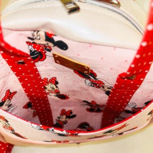 May include: A white handbag with red polka dot straps and trim. The interior features a pink fabric with a Minnie Mouse pattern. The bag has a zip closure and a small wooden tag.