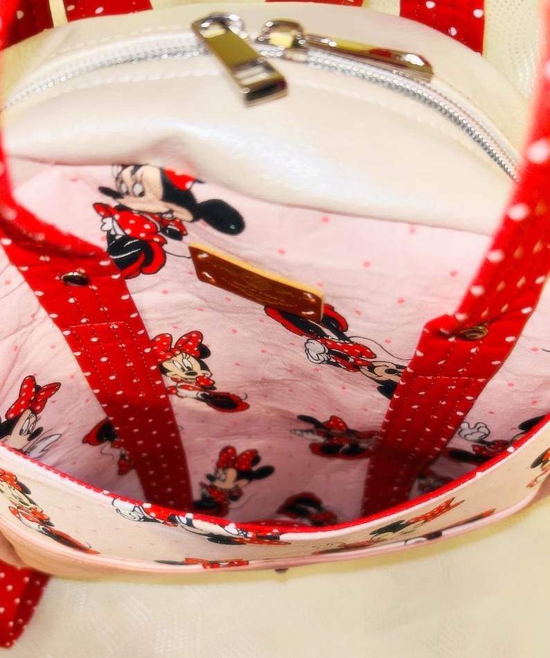 May include: A white handbag with red polka dot straps and trim. The interior features a pink fabric with a Minnie Mouse pattern. The bag has a zip closure and a small wooden tag.