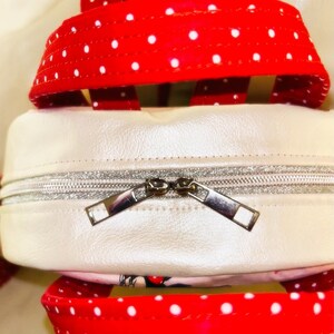 May include: Close-up of a white and red polka dot backpack. The backpack has a white body with a silver zip. The straps and top handle are red with white polka dots. The backpack is made of a shiny material.