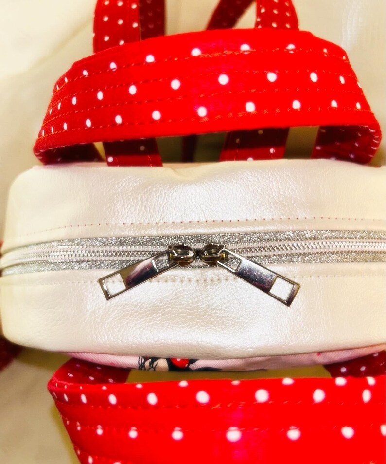 May include: Close-up of a white and red polka dot backpack. The backpack has a white body with a silver zip. The straps and top handle are red with white polka dots. The backpack is made of a shiny material.