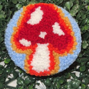 May include: A red and white mushroom with white spots, crocheted in a colorful yarn with blue, orange, and yellow accents. The mushroom is a handmade craft.