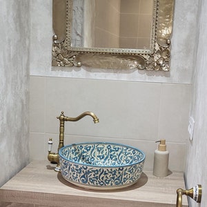 Op de afbeelding: Badkamermeubel met een decoratieve goudomrandde spiegel. De wastafel heeft een keramische wastafel met patroon, een messing kraan en een houten aanrechtblad. Daaronder een plank met opbergmanden en een handdoekenrek.