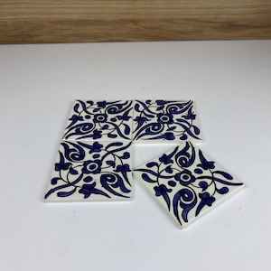 Peut inclure: Quatre dessous de verre carrés en céramique, blancs avec un motif floral bleu. Les dessous de verre sont disposés en carré, avec un légèrement décalé. Le motif présente des lignes tourbillonnantes et des motifs floraux.