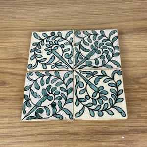Peut inclure: Quatre carreaux de céramique carrés avec un fond blanc et un motif de feuilles turquoise et noir. Les carreaux sont disposés ensemble, formant un plus grand carré. Le motif de feuilles est symétrique et détaillé.