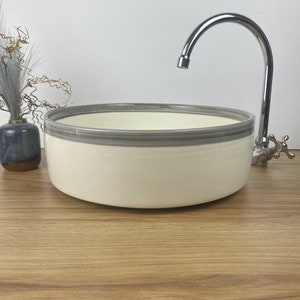 May include: A cream-coloured, round ceramic sink with a grey border, alongside a chrome tap. A small blue vase with decorative stems is to the left. The sink is on a wooden surface.