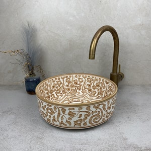 Op de afbeelding: Een witte keramische wastafel met een bruin bloemenpatroon. De wastafel is rond en heeft een lichte rand rond de rand. De wastafel staat op een grijs aanrecht.