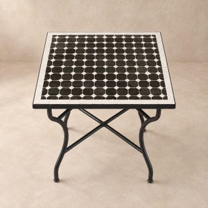 May include: A square table featuring a black and white mosaic tile top with a repeating octagonal pattern. The table is supported by a black metal frame with four curved legs connected by a central X-shaped cross brace for structural stability.