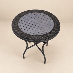 Peut inclure: Une table de bistrot ronde avec un plateau en mosaïque composé de carreaux octogonaux gris clair et de petits accents carrés noirs, entouré d'une bordure sombre de carreaux noirs. La table possède un cadre en métal noir à quatre pieds incurvés.