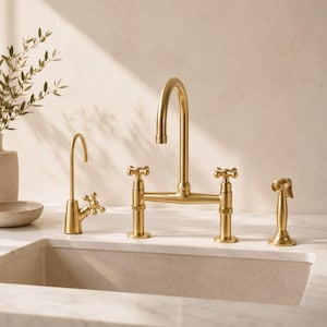 Può includere: Un set di rubinetti da cucina in oro spazzolato composto da un rubinetto a ponte ad arco alto con due manopole a croce, un rubinetto separato più piccolo per acqua filtrata con manopola abbinata e una doccetta laterale, su un piano in marmo.