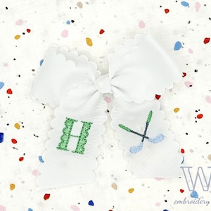 May include: White scalloped bow with green embroidered letter H and blue and green embroidered golf clubs. The bow is on a white background with colorful confetti.