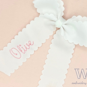 May include: White scalloped ribbon with the name "Olivia" embroidered in pink thread. The ribbon is tied into a bow.
