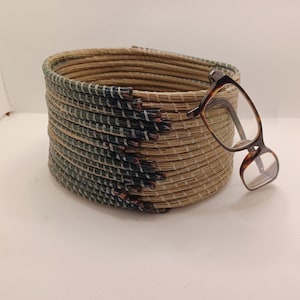 May include: A round woven basket with a natural tan and green color scheme. The basket has a textured appearance and is decorated with a dark blue and brown pattern. A pair of tortoiseshell eyeglasses rests on the side of the basket.