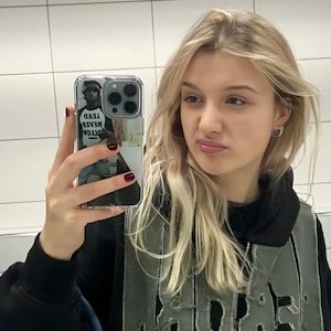 Op de afbeelding: Een persoon houdt een transparante telefoonhoes met een zwart-witfoto van een persoon die een shirt draagt met de tekst "OTTOM WEAR MEAD TEAD".