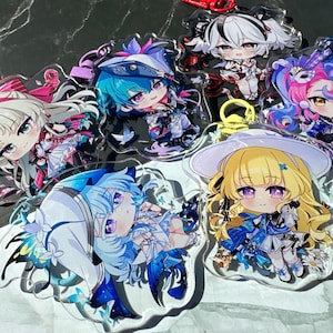 May include: A selection of anime-style acrylic keyrings, each depicting a character with large eyes and detailed outfits. The keyrings feature diverse hairstyles and vibrant colours, including blue, pink, and yellow. Each keyring has a metal clasp.