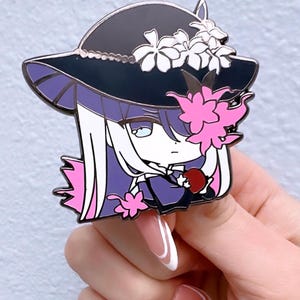 May include: Enamel pin depicting an anime-style character with long white hair, a black hat decorated with white flowers, and pink floral details. The character holds a red apple. The pin has a purple and black colour scheme.