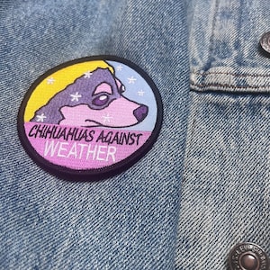 May include: A purple and pink embroidered patch with a cartoon chihuahua wearing a yellow hat. The patch says "Chihuahuas Against Weather" in black text.