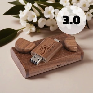 Może przedstawiać: Drewniany pendrive z wygrawerowanym tekstem "Hannelie Bloem fotografie". Pendrive umieszczony jest na drewnianym pudełku z dwoma drewnianymi akcentami. Tło obejmuje białe kwiaty i numer "3.0".