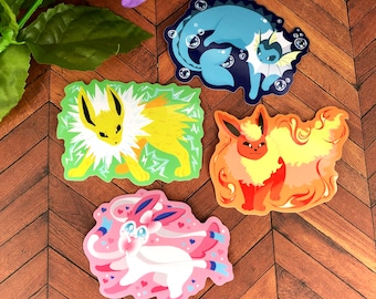 May include: Four colorful stickers featuring different Pokemon characters. The stickers are on a wooden surface. The characters are a yellow and white electric type Pokemon, a blue and white water type Pokemon, a red and orange fire type Pokemon, and a pink and white fairy type Pokemon.