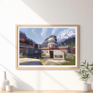 Op de afbeelding: Een ingelijste print van een videogame-omgeving met een basis met rode en witte accenten, zeecontainers en een berglandschap. De print zit in een licht houten frame.