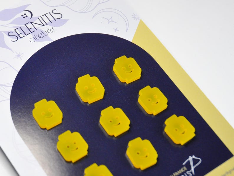 May include: Eight yellow resin buttons shaped like Lego minifigures with smiling faces. The buttons are arranged in a circle on a dark blue background.