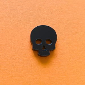 Könnte beinhalten: Ein schwarzer Totenkopf-Anhänger oder -Charm. Der Totenkopf ist aus glattem, massivem Material gefertigt und hat ein schlichtes, klassisches Design.