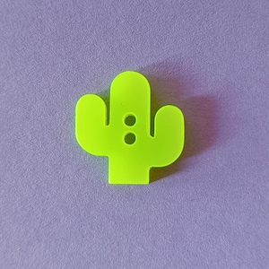 Op de afbeelding: Een heldere, neon-groene cactusvormige knoop met twee kleine gaatjes, tegen een zachte paarse achtergrond. Het ontwerp van de knoop bootst een gestileerde cactusplant na, ideaal voor naai- of knutselprojecten.