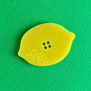 May include: A bright yellow lemon-shaped button with four small black holes and a pattern of small dots. The button is set against a vibrant green background, creating a striking contrast. This button is ideal for sewing projects.