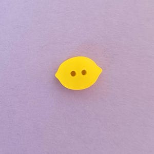 Könnte beinhalten: Ein leuchtend gelber, zitronenförmiger Knopf mit zwei Löchern, vor einem weichen, hellvioletten Hintergrund. Dieser dekorative Knopf ist ideal für Nähprojekte und verleiht Kleidung oder Bastelarbeiten eine fröhliche Note.