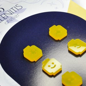 May include: Six yellow plastic buttons with a smiley face design. The buttons are shaped like a person's head and have a raised smiley face on the front. The buttons are arranged in a semi-circle on a dark blue surface.