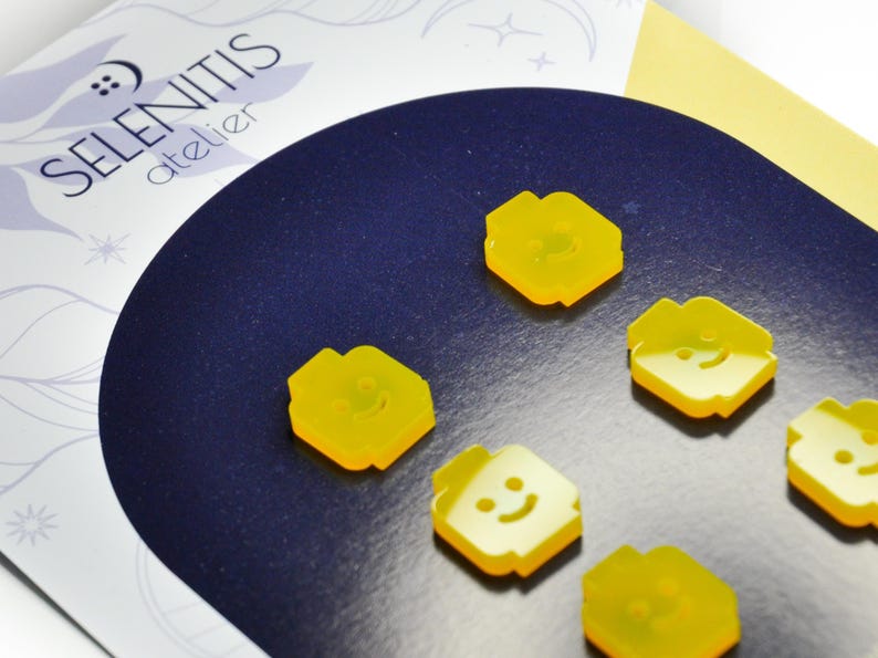 May include: Six yellow plastic buttons with a smiley face design. The buttons are shaped like a person's head and have a raised smiley face on the front. The buttons are arranged in a semi-circle on a dark blue surface.